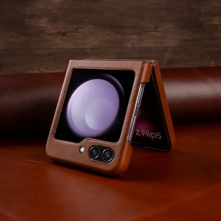 A Samsung Galaxy Z Flip5 smartphone in a genuine leather case is partially folded and resting on a brown surface. The outer display shows a circular graphic, and the dual rear cameras are visible for stylish shockproof protection.