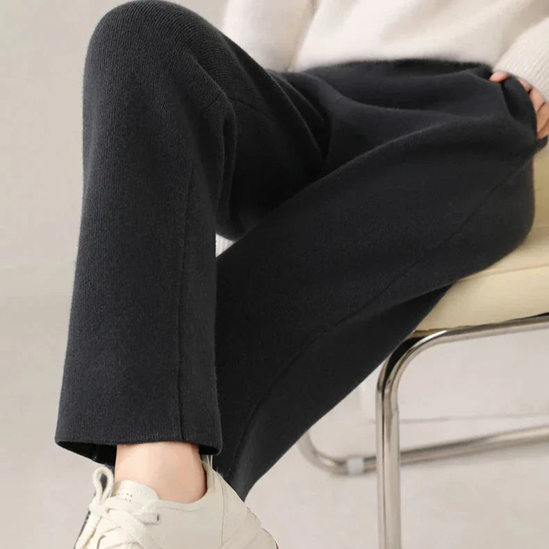 A person seated on a chair, wearing black pants with a slim tailored leg, a light-colored sweater, and white sneakers. The photo focuses on their legs and part of their hand resting on their lap.