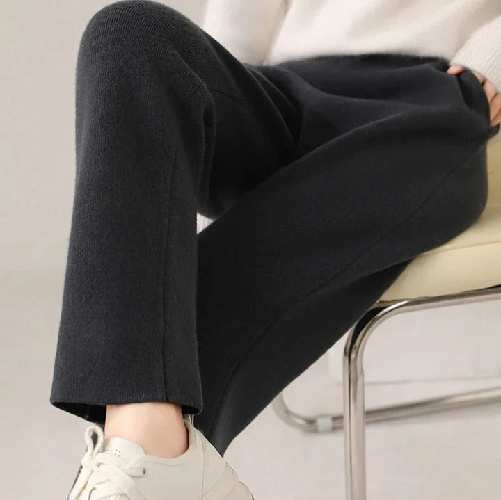 A person seated on a chair, wearing black pants with a slim tailored leg, a light-colored sweater, and white sneakers. The photo focuses on their legs and part of their hand resting on their lap.