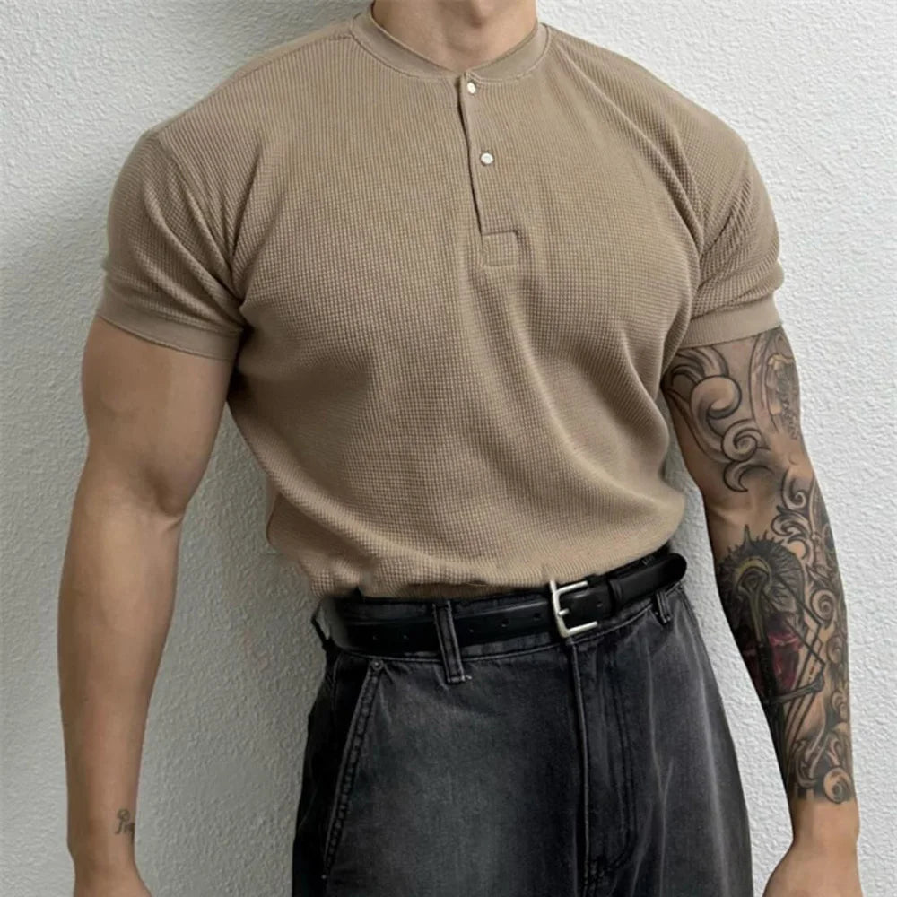 A person wearing a beige, form-fitting ribbed Henley, black belt, and dark jeans stands against a light textured wall. Their right arm is heavily tattooed, and their face is not visible in the image.
