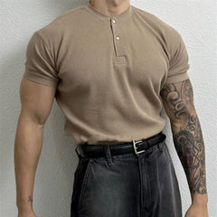A person wearing a beige, form-fitting ribbed Henley, black belt, and dark jeans stands against a light textured wall. Their right arm is heavily tattooed, and their face is not visible in the image.