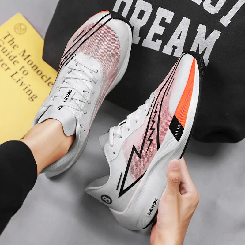 A person wearing white and pink running shoes with black and orange accents holds one shoe in hand. Featuring a shock-absorbing sole, these shoes also display text and graphics. A yellow book and black sweatshirt are in the background.