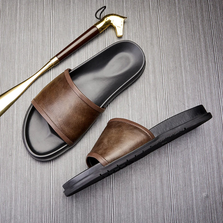 A pair of brown and black handmade leather sandals rests on a textured gray surface, next to a gold and brown shoehorn.