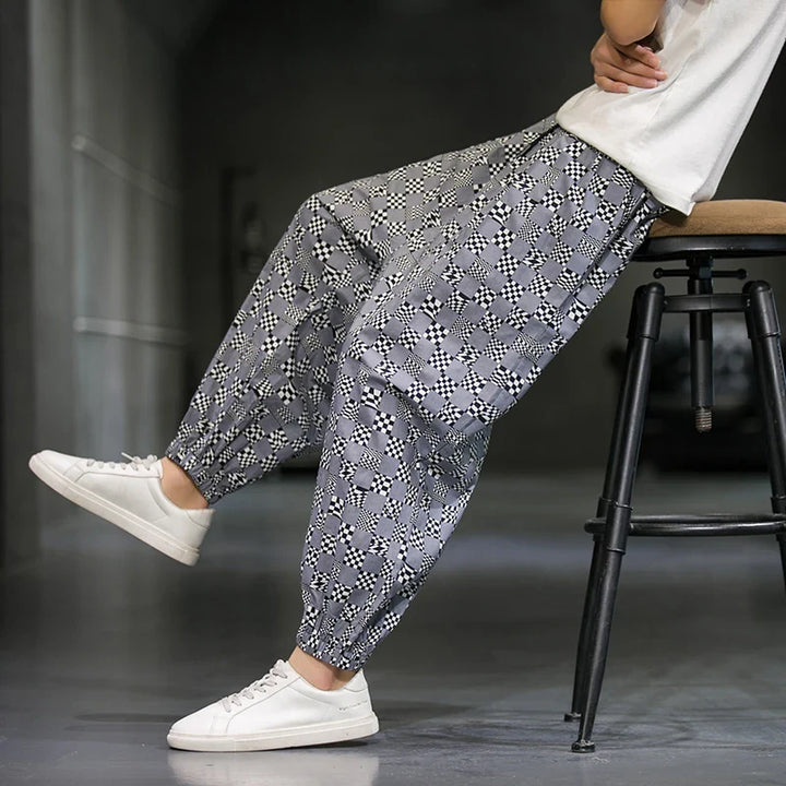 A person wearing patterned gray cotton joggers and white sneakers sits sideways on a tall stool in a minimal, modern indoor space. Only their lower body is visible, making a subtle fashion statement.