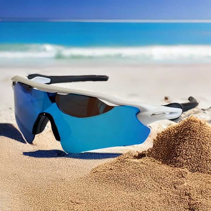 A pair of blue mirrored Polarized Cycling Sunglasses sits on sandy beach with a small mound of sand, as turquoise ocean waves and a clear blue sky form the perfect backdrop.