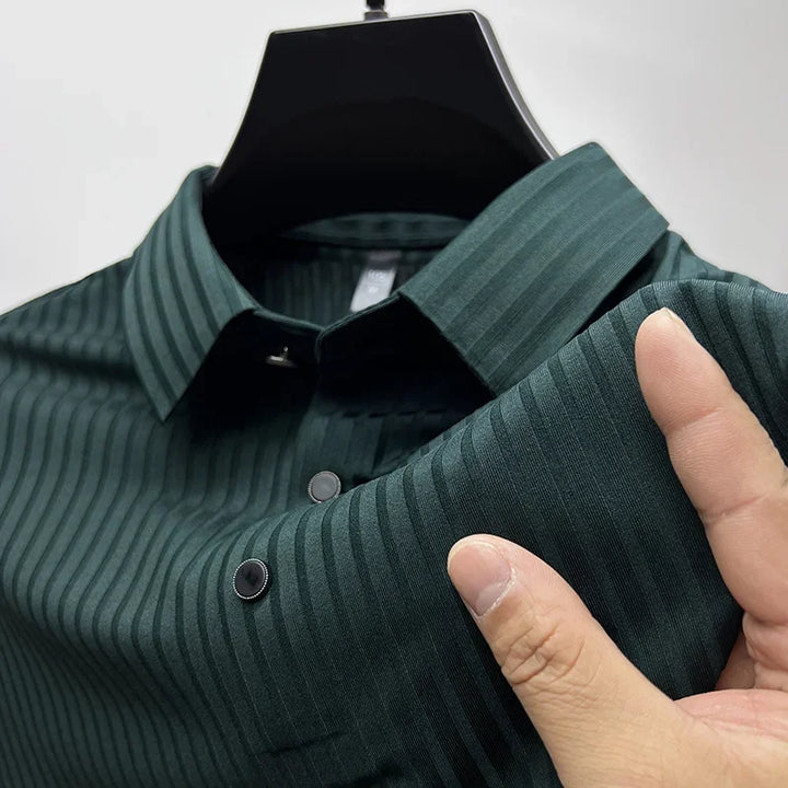 A close-up of a dark green, vertically striped dress shirt on a hanger, with a hand holding and slightly pulling the luxurious silk fabric to display its texture and pattern.
