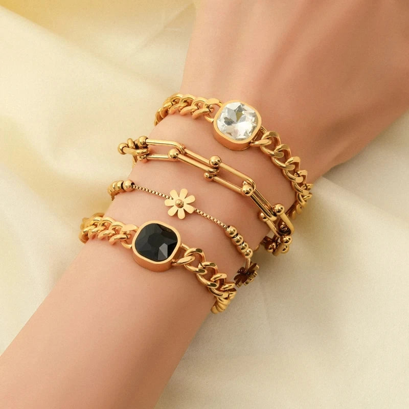 A hand wearing four gold bracelets, crafted from 316L stainless steel with unique designs—a large clear gem, a black gem, a flower charm, and small gold beads—rests on light cream fabric. Each piece features an adjustable clasp for a perfect fit.