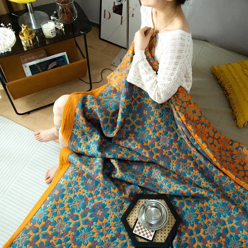 A woman sits on a bed holding a vibrant ethnic pattern blanket in blue and orange hues. Next to her is a small table with a silver tray, cups, and a magazine. The room is cozy with soft lighting and modern decor.
