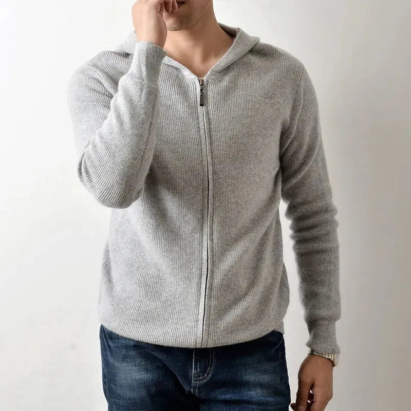 A person wearing a light gray cashmere hoodie and blue jeans stands against a plain white background, with one hand partially covering their mouth, showcasing the luxurious cashmeres lightweight insulating comfort.