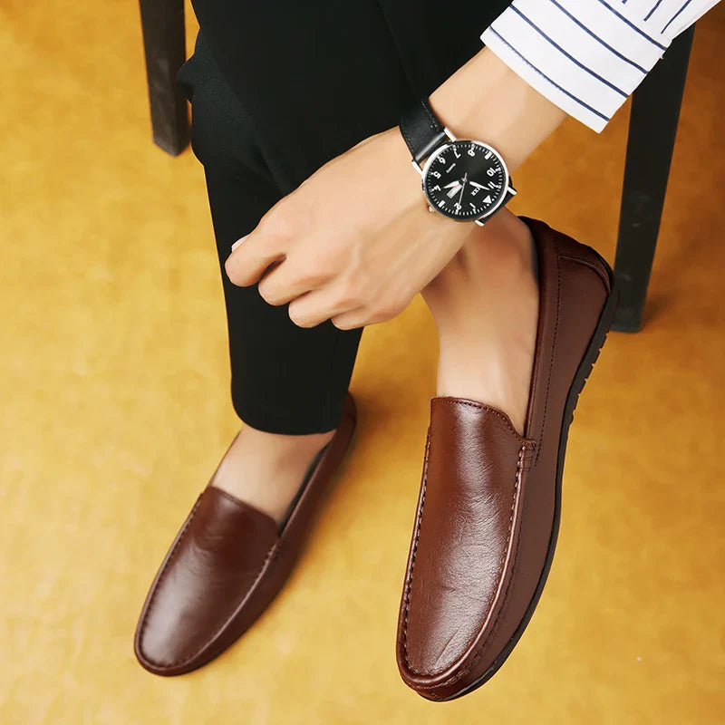 A person wearing black pants and a striped shirt sits on a chair, showing off luxury shoes—brown leather loafers with a cushioned footbed—and a black wristwatch with a round face. The floor is light brown.