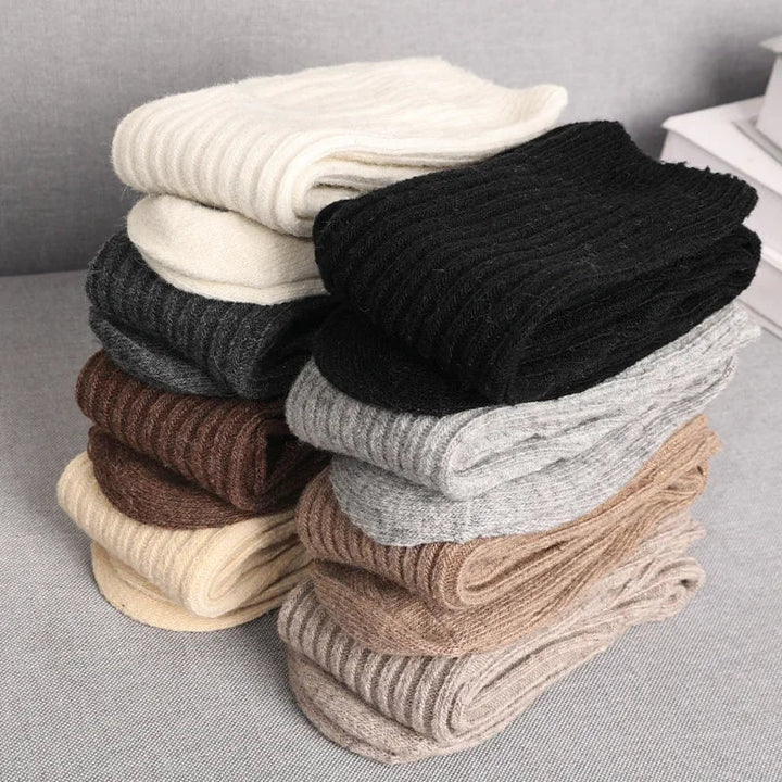 Two stacks of folded, ribbed knit socks in various neutral colors, made from a Cashmere cotton blend for superior insulation, placed on a light gray surface.