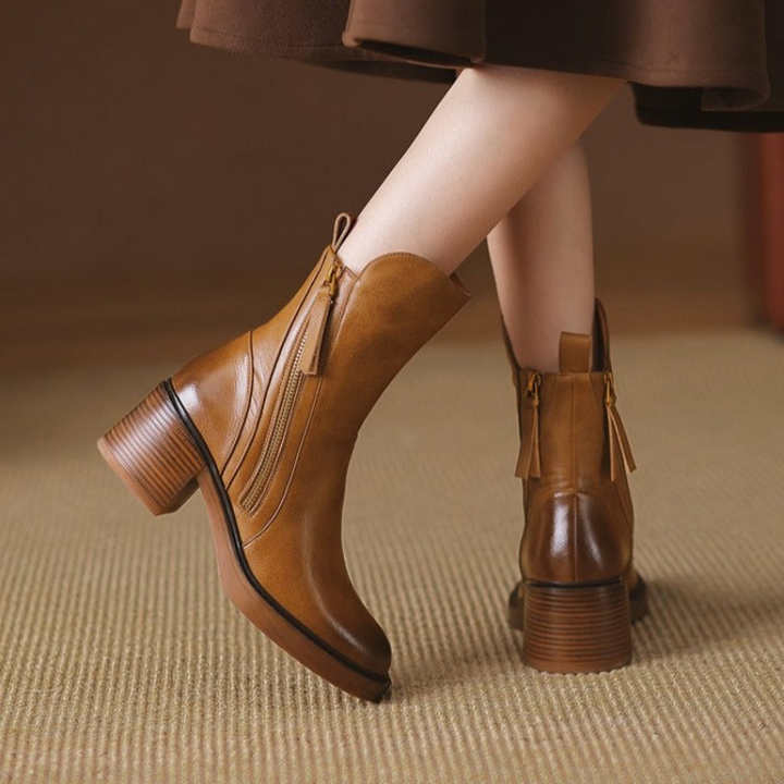 A person wearing brown heeled ankle boots with cushioned insoles and tassel details stands on a beige carpet; the polished leather boots perfectly complement a brown skirt.