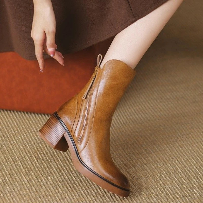 A person wearing a brown dress sits with one leg crossed, showing off a polished leather ankle boot with a side zipper and chunky heel, featuring a cushioned insole, on a beige textured carpet.
