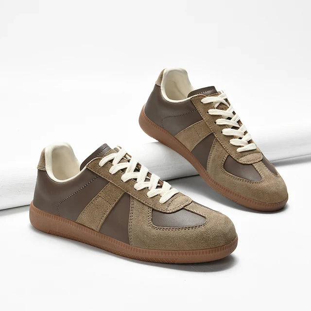 A pair of stylish brown and beige lightweight breathable sneakers with white laces, tan rubber soles, and premium cow suede uppers are displayed on a white surface, with one shoe propped against a white cylindrical object.