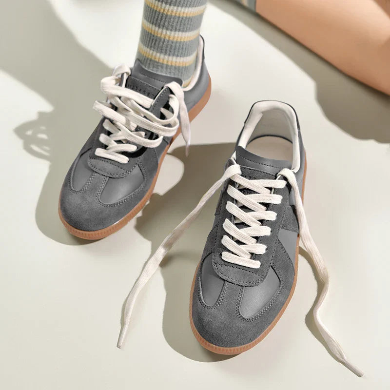 A pair of gray and white premium cow suede sneakers with gum soles rest on a light surface; one shoe is lying flat while the other stands upright. A person wearing striped socks is partially visible in the background.