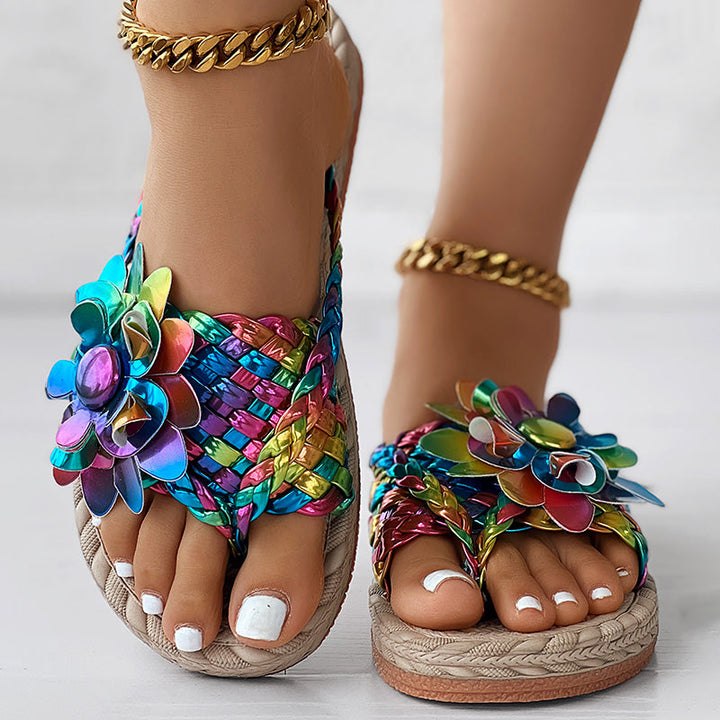 Close-up of feet wearing colorful, woven sandals with shiny flower decorations—perfect vacation essentials for women's fashion casual shoes—white toenail polish and gold anklets complete the look, standing on a light surface.