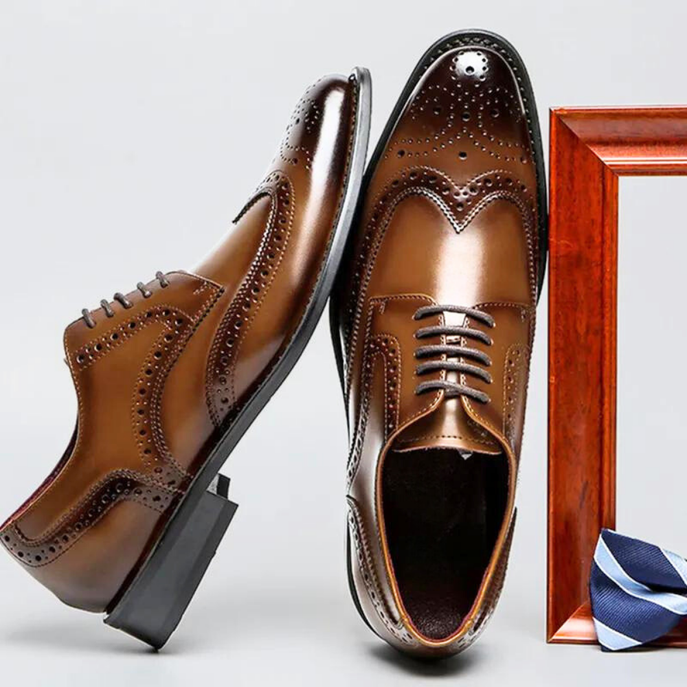 A pair of shiny brown premium leather brogue dress shoes, one lying sideways and the other upright, displayed next to a wooden picture frame and a small, folded blue patterned cloth. Features a cushioned insole for added comfort.