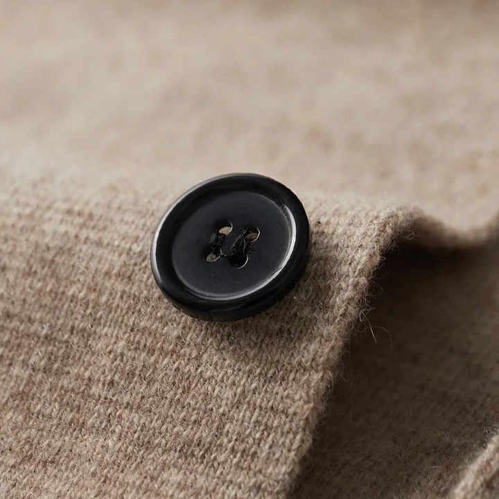 A close-up of a black, four-hole button sewn onto a textured, light brown fabric, likely part of a tailored cardigan. The button sits near the edge of the soft, slightly woolly material.