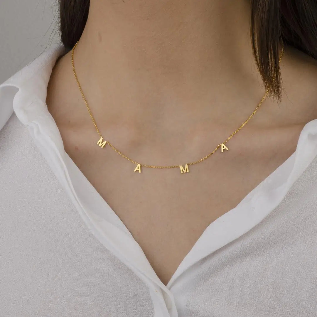 A woman wearing a white shirt and a gold Mothers Day necklace with letter charms spelling MAMA, showcasing stylish minimalist jewelry.
