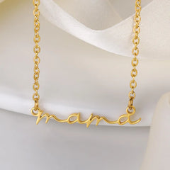 A gold-plated mama necklace with a delicate chain features a pendant that spells “mama” in cursive script, displayed against a soft, cream-colored fabric background.