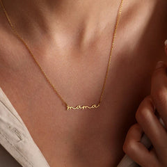 A close-up of a person wearing a delicate gold-plated mama necklace in cursive script. The Mothers Day necklace rests on their collarbone, paired with a light-colored top.