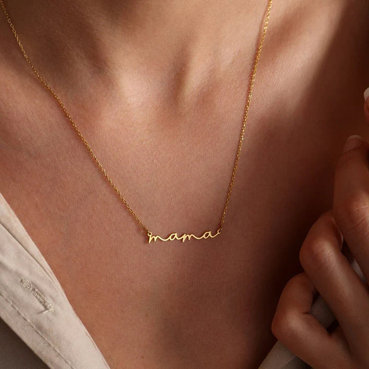 A close-up of a person wearing a delicate gold-plated mama necklace in cursive script. The Mothers Day necklace rests on their collarbone, paired with a light-colored top.
