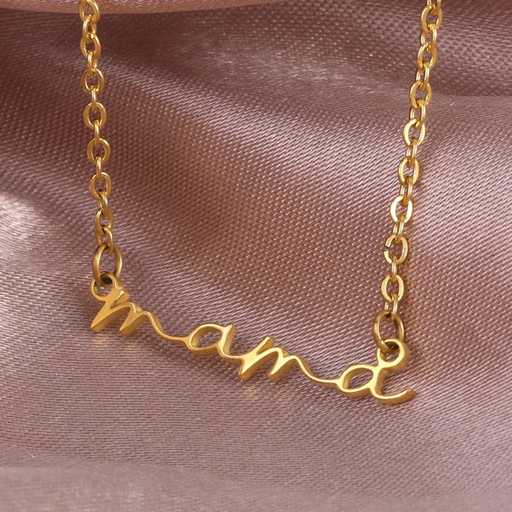 A gold-plated mama necklace with the word mama in cursive script, displayed on a soft, light pink satin fabric background—perfect as a thoughtful Mothers Day necklace.