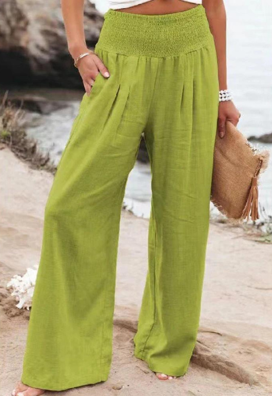 A person stands outdoors near rocks and water, wearing wide-leg, high-waisted lime green summer pants and holding a woven clutch purse with tassels. Only the lower half of the body is visible.