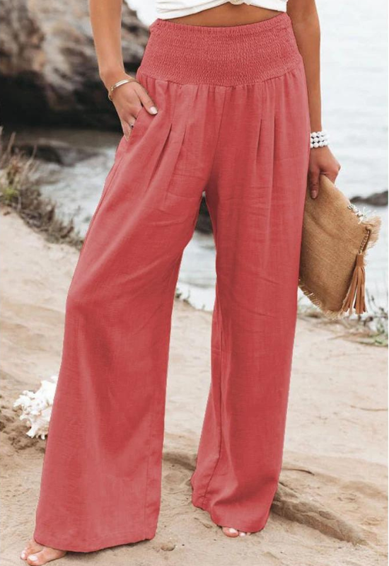 A person stands outdoors in wide-leg, high waist coral pink summer pants, holding a tan clutch bag and wearing a white shirred top with a beaded bracelet, against a backdrop of rocky shore and sparkling water.