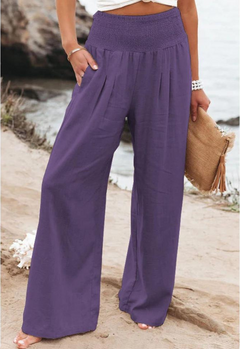 A person stands outdoors near a rocky shoreline, wearing loose, high waist summer pants in purple and holding a woven clutch with tassels. Only the lower body is visible.