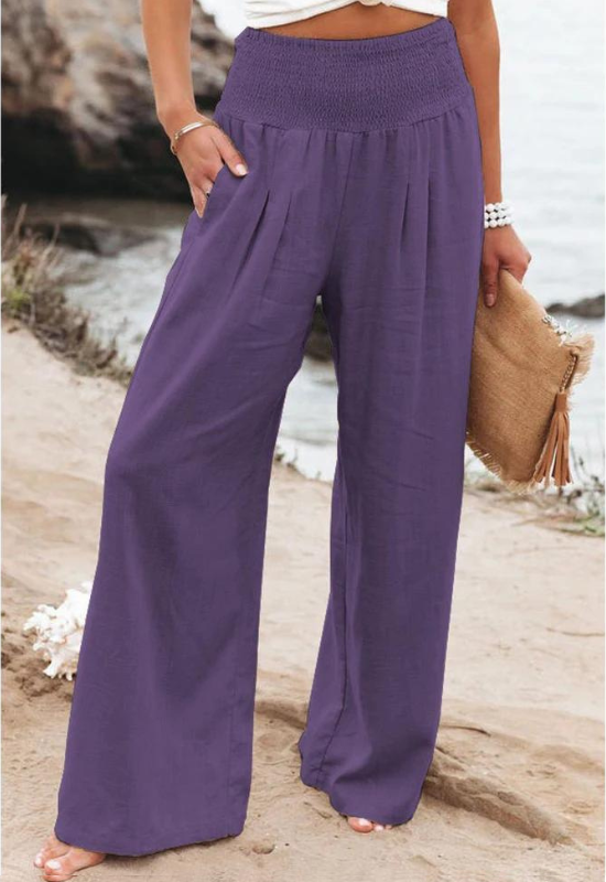 A person stands outdoors near a rocky shoreline, wearing loose, high waist summer pants in purple and holding a woven clutch with tassels. Only the lower body is visible.