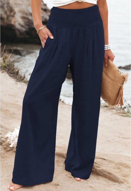 A person stands outdoors near the shore, wearing navy blue high waist wide-leg summer pants and holding a tan clutch with tassels. Only the lower half of the body is visible.