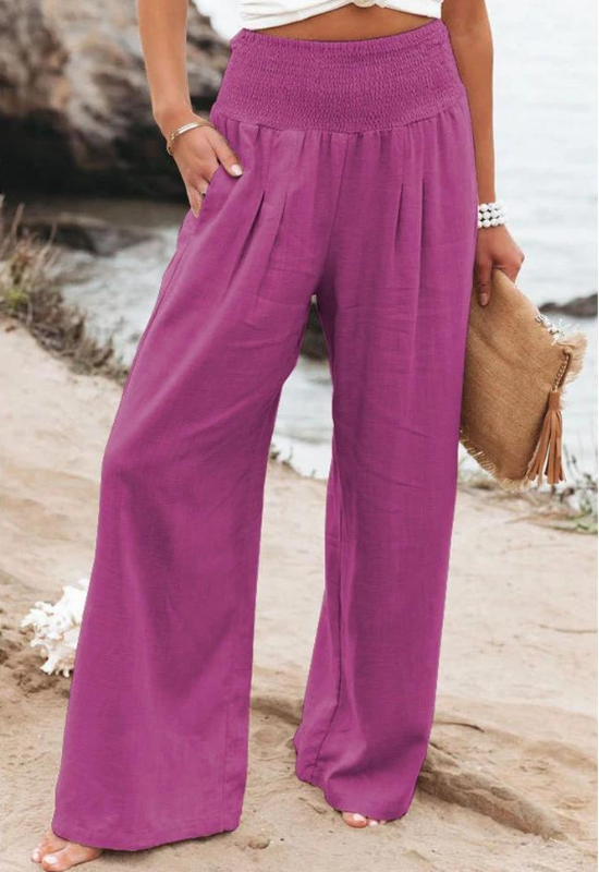 A person stands outdoors near water, wearing high waist, wide-leg purple summer pants, a white top, white beaded bracelets, and holding a tan woven clutch with tassels.