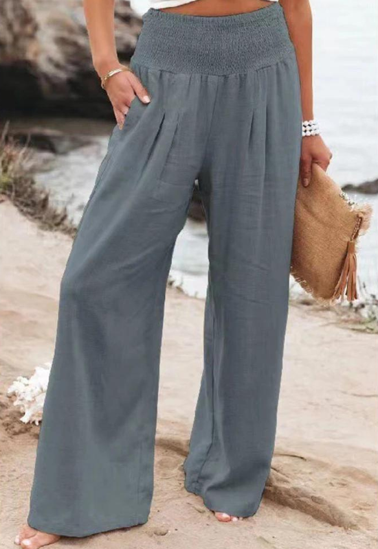 A person standing outdoors by the water, wearing loose-fitting, high waist summer pants and a white or light top, holding a woven clutch bag with tassels. Only the lower half of their body is visible.