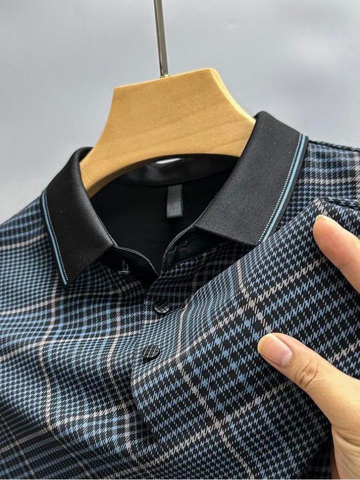 A hand holds a plaid-patterned, temperature-regulating shirt with a black collar on a wooden hanger. This breathable shirt features a mix of blue, black, and white colors, set against a neutral background.