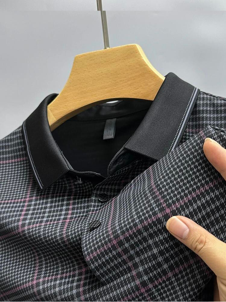 A close-up of a black collared shirt with a pink and white plaid pattern, displayed on a wooden hanger. A hand holds the sleeve, highlighting the breathable fabric texture and comfortable design.