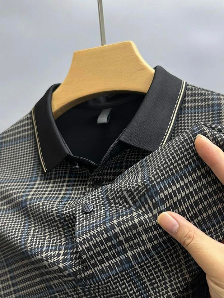 A hand holds a breathable, checkered collared shirt with a dark collar on a wooden hanger against a plain background. The shirt features a black, white, and blue plaid pattern, perfect for staying comfortable all day.