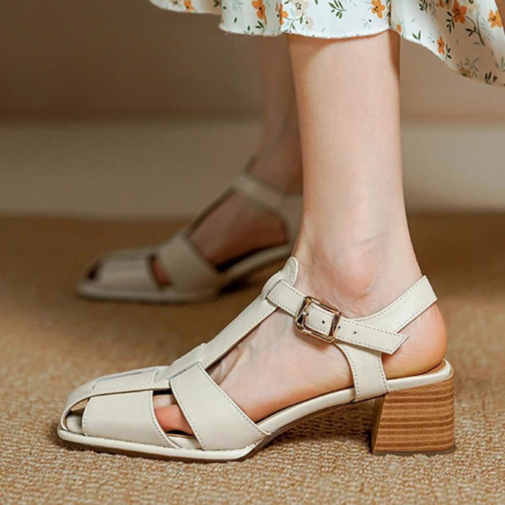 A person wearing handcrafted, beige, open-toe strappy sandals with a medium block heel and buckle. The sandals, paired with a light floral dress, are made from Genuine Italian Leather and rest on a tan carpet.