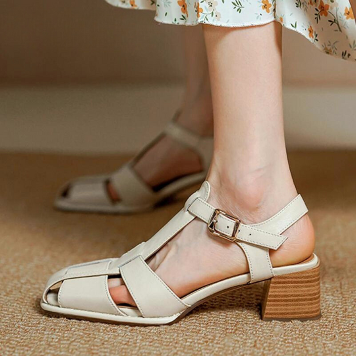 A person wearing handcrafted, beige, open-toe strappy sandals with a medium block heel and buckle. The sandals, paired with a light floral dress, are made from Genuine Italian Leather and rest on a tan carpet.