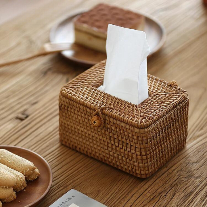 A handcrafted rattan tissue box with a button closure sits on a wooden table. In the background, plates with pastries and biscuits add to the cozy, inviting ambiance and showcase charming organic textural decor.