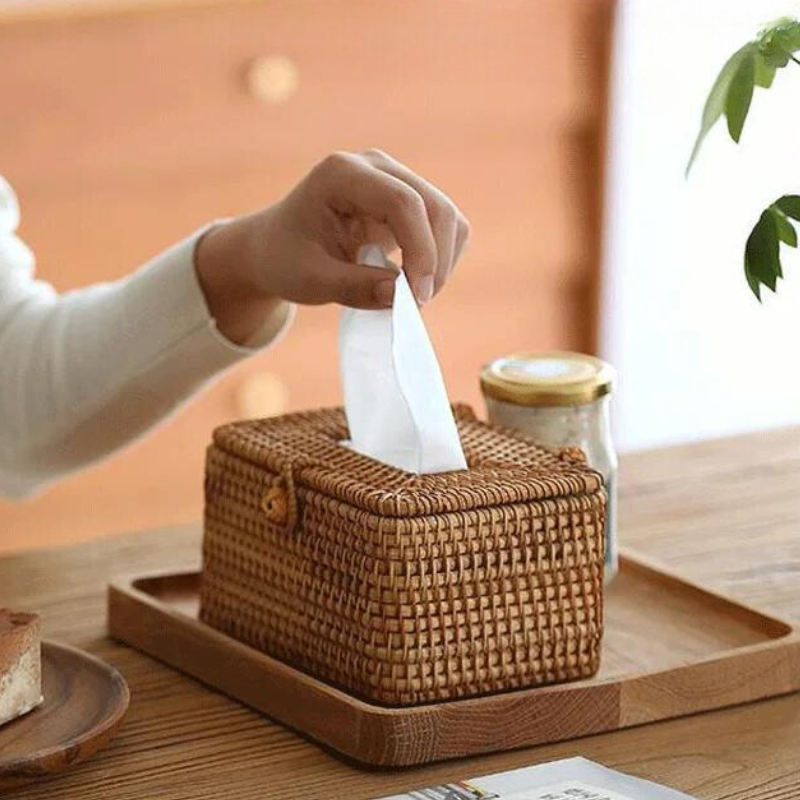 A hand pulls a tissue from a handcrafted rattan tissue box on a wooden tray, with a jar and a slice of cake nearby on the table—perfect for those who appreciate eco-friendly materials and organic textural decor.