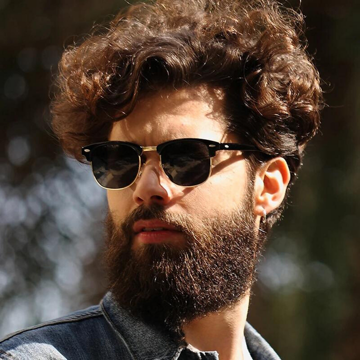 A man with curly brown hair, a full beard, and mustache wears vintage sunglasses with UV protection and a denim jacket outdoors. The background is blurred with hints of greenery and sunlight.