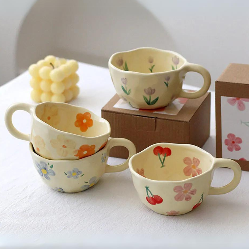 Four vintage teacups with pastel floral and fruit patterns sit on a white table. Two cups are stacked, one rests on a box, and a decorative candle shaped like grapes is in the background.