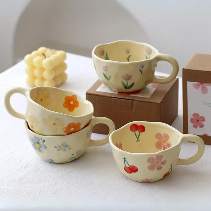 Four vintage teacups with pastel floral and fruit patterns sit on a white table. Two cups are stacked, one rests on a box, and a decorative candle shaped like grapes is in the background.