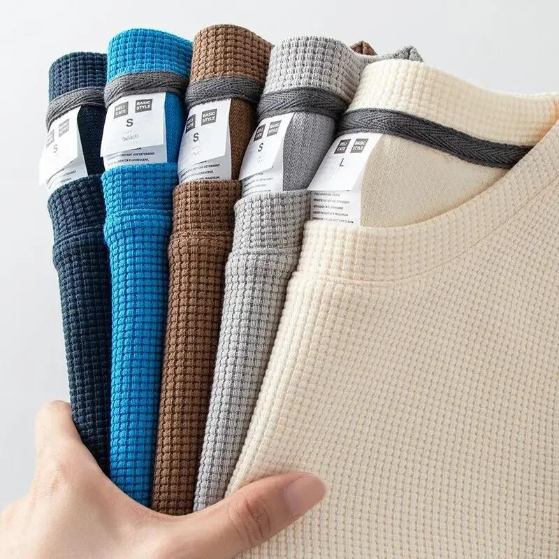 A hand holds five folded waffle knit shirts in blue, brown, gray, and cream—each made from high-quality cotton and featuring a size tag near the collar.