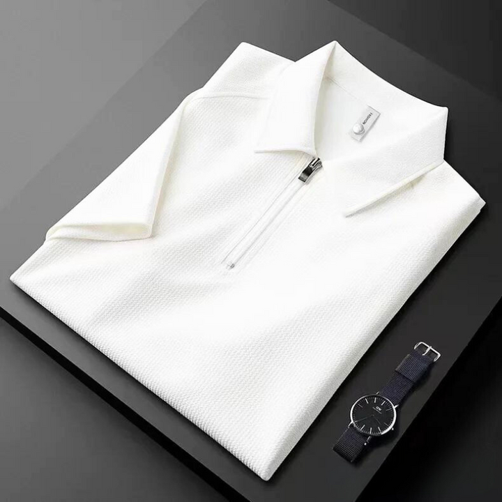 A neatly folded white mens silk polo shirt with a front zipper is placed on a black surface next to a minimalist black wristwatch with a black strap.