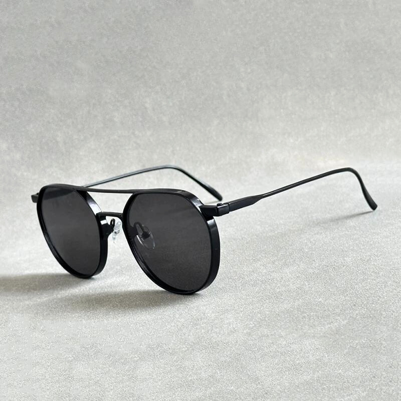 Black round sunglasses with thin, lightweight alloy frames and dark lenses, placed on a light gray textured surface. The arms are extended, highlighting their minimal, modern style and offering both comfort and UV400 protection.