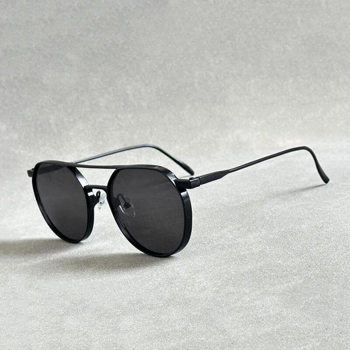 Black round sunglasses with thin, lightweight alloy frames and dark lenses, placed on a light gray textured surface. The arms are extended, highlighting their minimal, modern style and offering both comfort and UV400 protection.