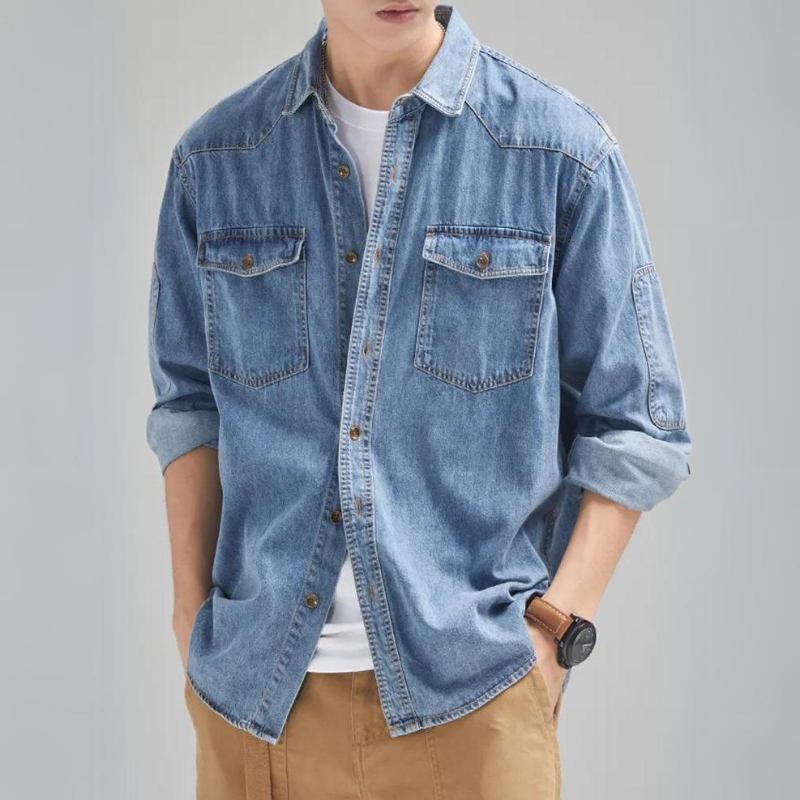 A person wearing a rugged denim shirt over a white t-shirt, with rolled-up sleeves, tan pants, and a black wristwatch, standing with hands in pockets.