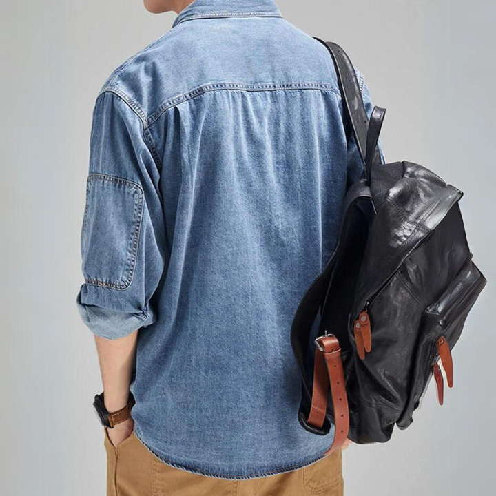 A person wearing a rugged denim shirt and khaki pants is seen from behind, carrying a black leather backpack with brown straps over their right shoulder.
